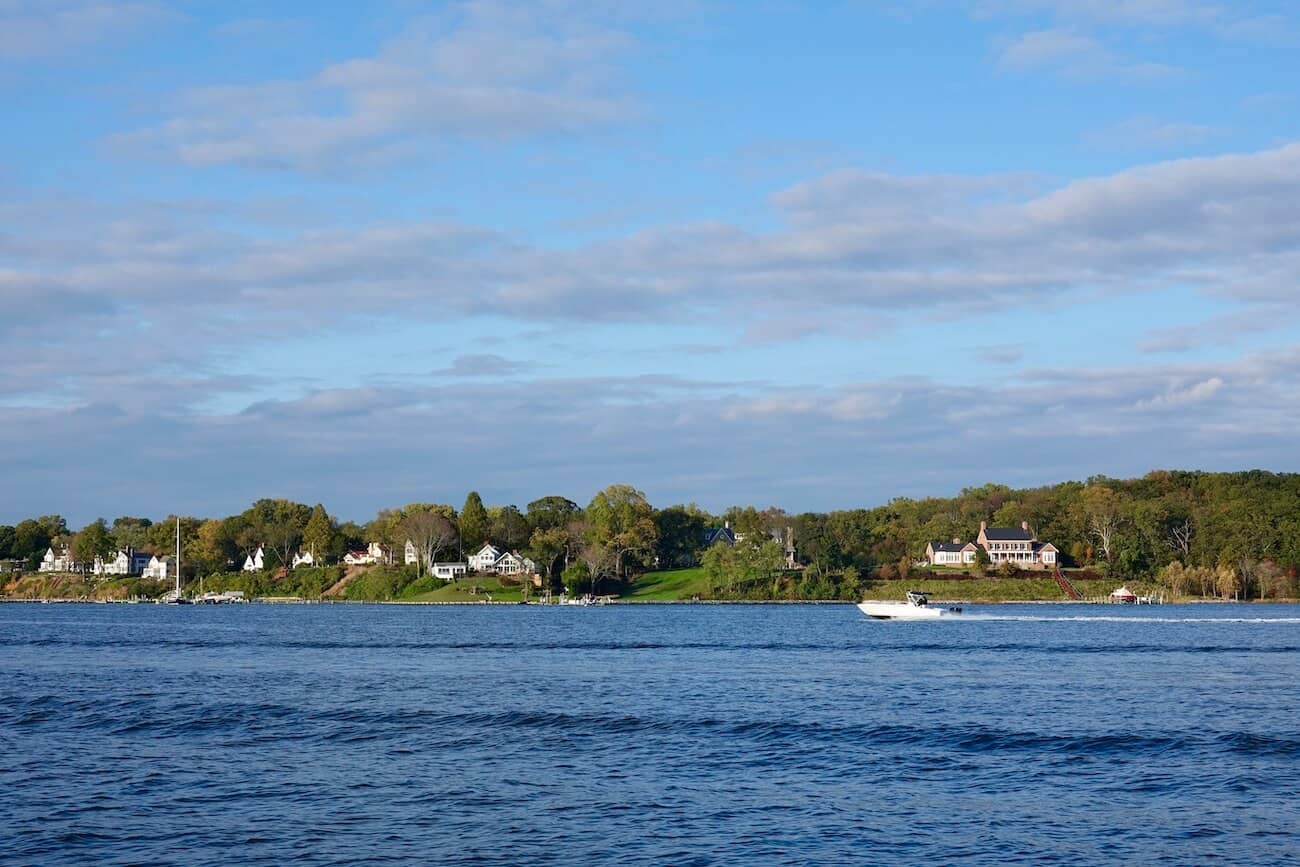 A boat speeds by on the water with homes along the shore in the distance.
