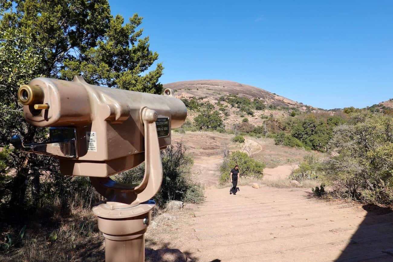 A telescope looks towards the dome of Enchanted Rock as a hiker walks forward.
