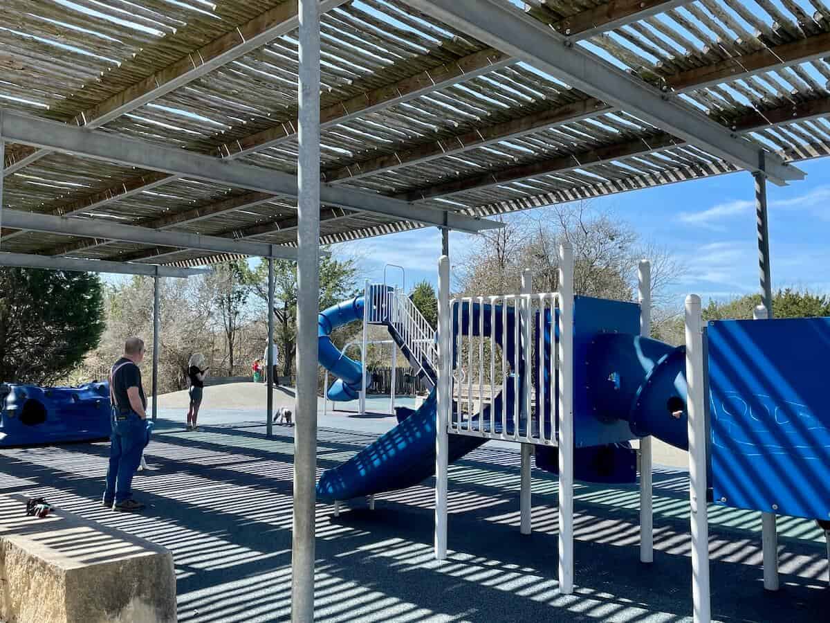 An outdoor playground with blue play structures sit under a pergola.