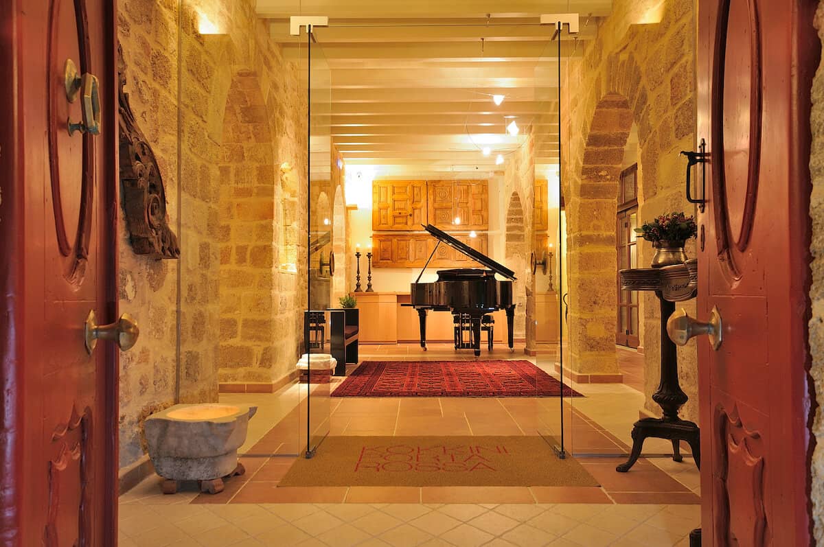 A grand piano sits in the center of a stone foyer just beyond red entrance doors.