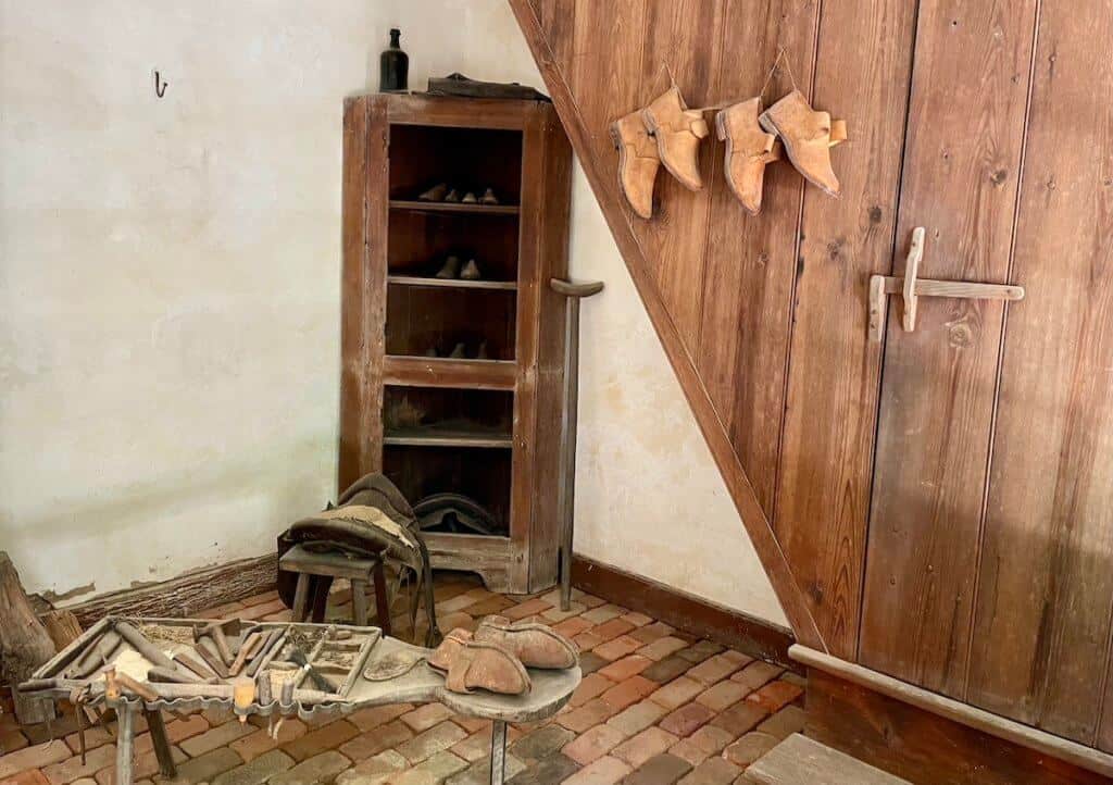 Tools and materials for making shoes are displayed on a wooden wall and brick floor.