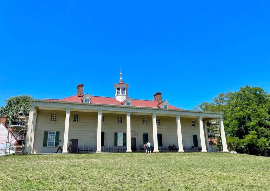 Eight pillars form a porch on aa two story house with a red roof and cupola.