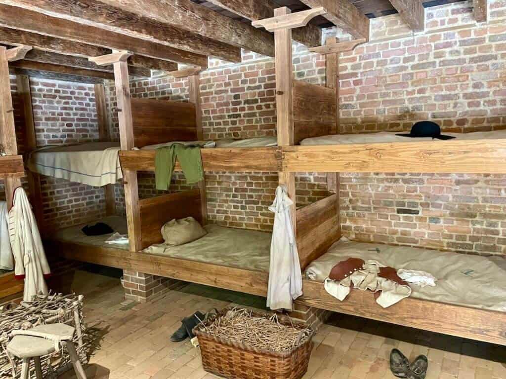 Wooden bunk beds are lined up on a brick floor with garments and shoes.