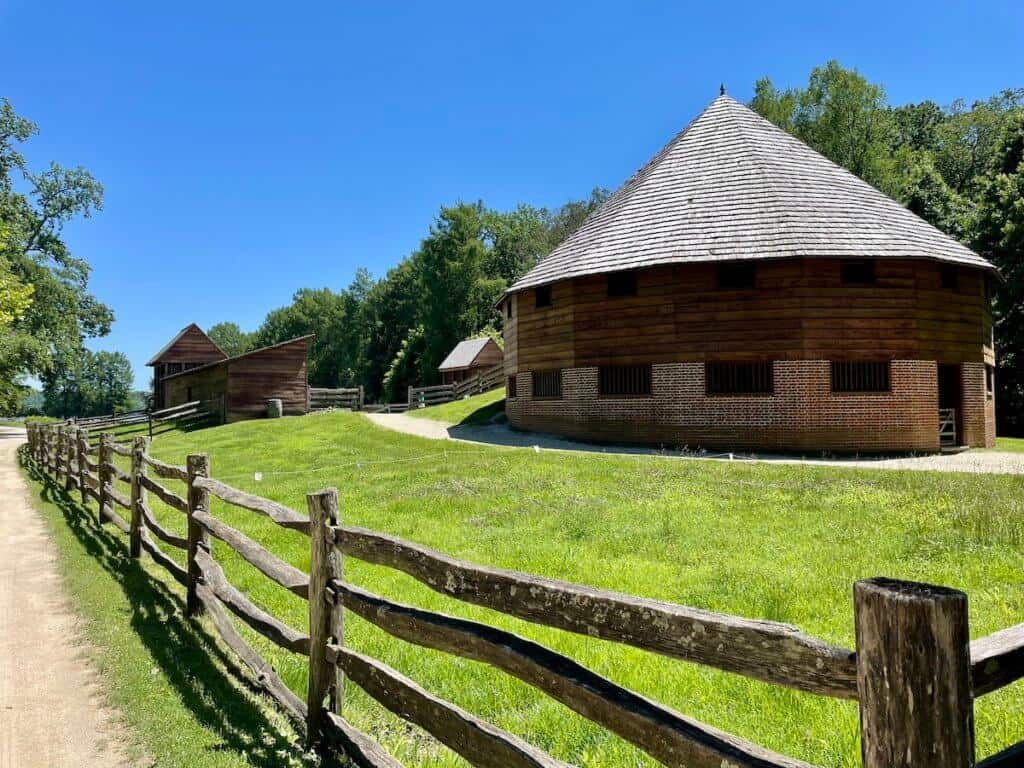 A large brick and wooden barn sits in a grassy area surrounded by an old wooden stockade fence.