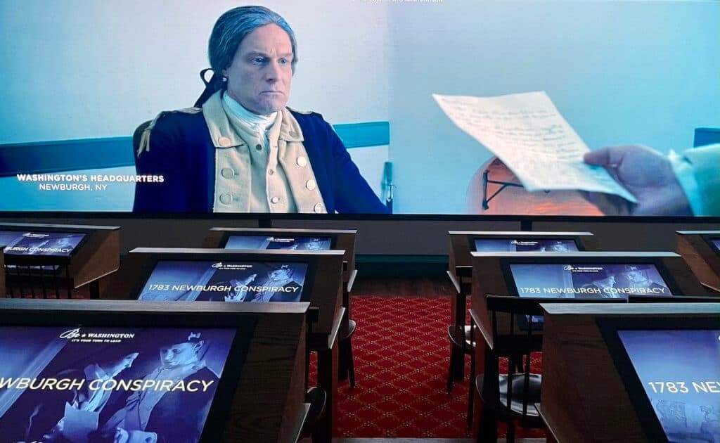 A large screen displays a video; desks sit in front with computer screens for visitors to interact with the video.