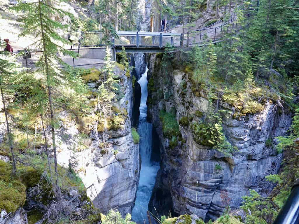 A river spills out between crevices in a limestone canyon in Canada; people stand on a footbridge above it.