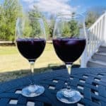 Two wine glasses hold red sangria on a black table in the foreground with deck stairs and white pines stand in the background..