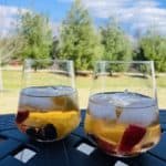 Two glasses of white sangria sit on a black table outside with white pine trees stand in the background.