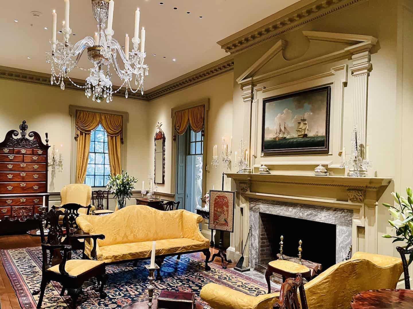 Formal Queen Anne furniture is placed on an oriental carpet under a glass chandelier in a painted paneled room with a fireplace.