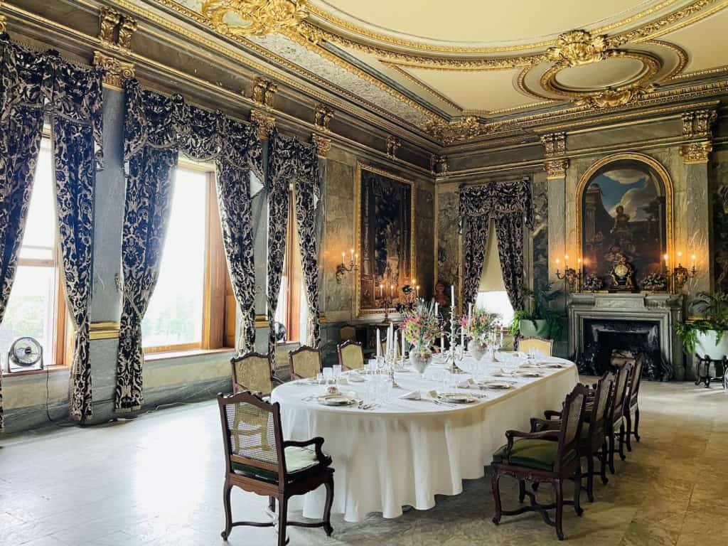 A large dining table with a white tablecloth is set for 10 people in a large room with elaborate gold scrollwork on the ceiling and tall windows draped with blue and white curtains.