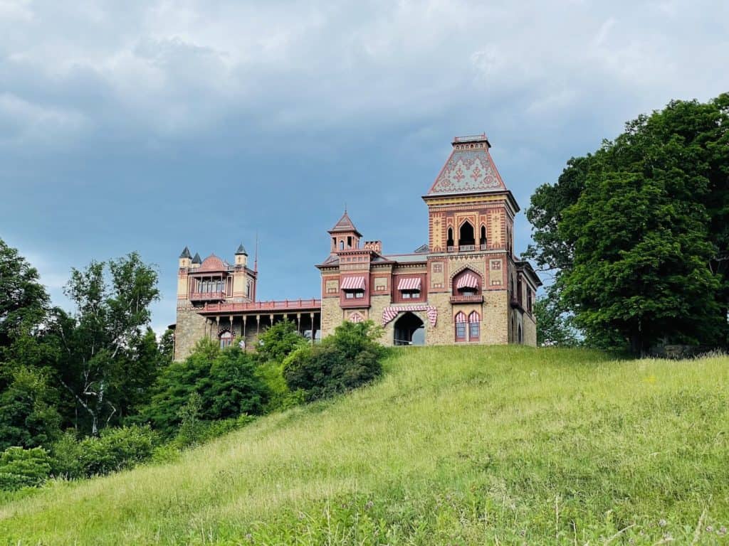 A large multi-colored stone and tiled home sits atop a grassy hill.