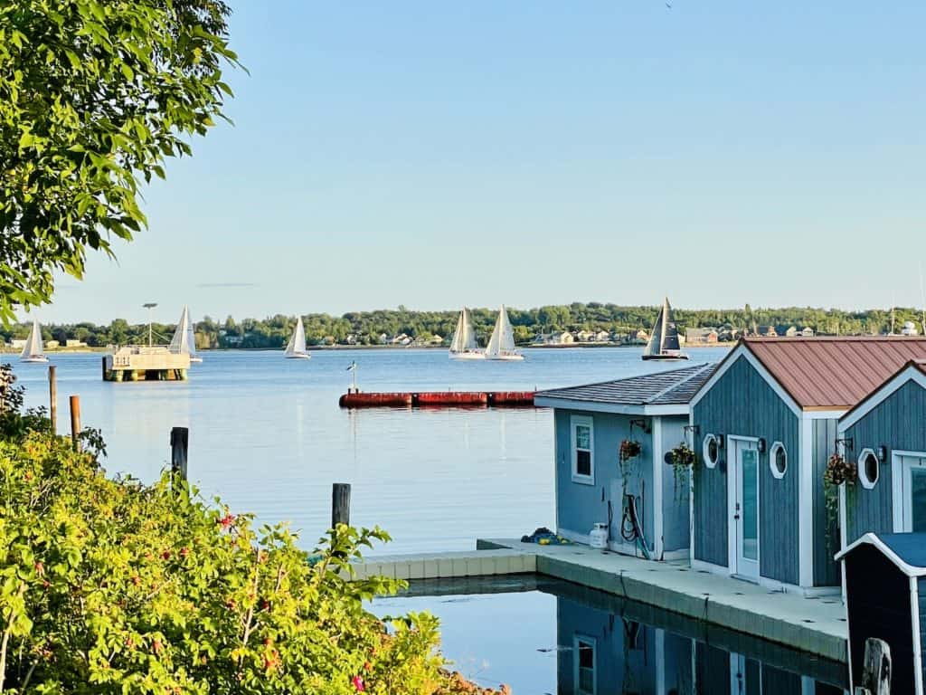 Small blue sheds stand along a boardwalk in a marina while sailboats glide on the water in the harbor in Charlottetown, PEI.