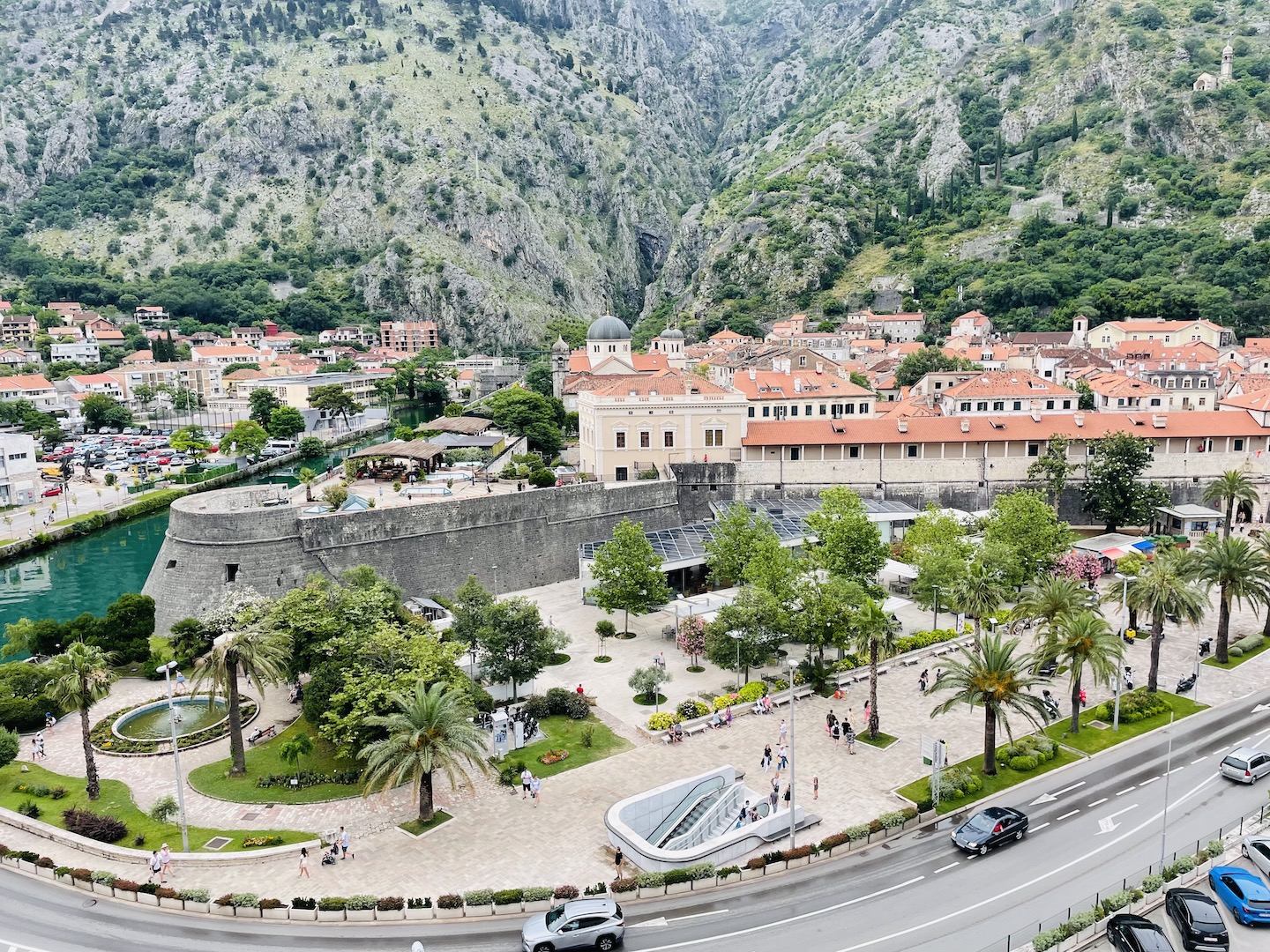 The medieval Kotor's walled city is surrounded by mountains and a paved promenade with palm trees.