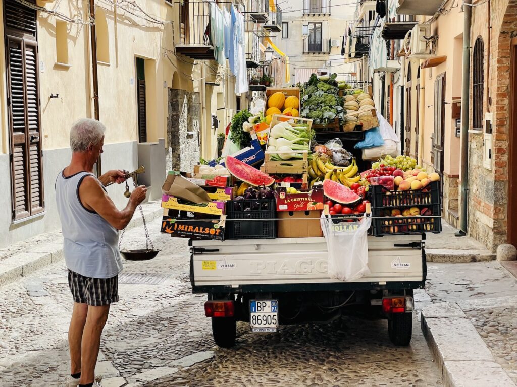 A man weighs some fruit from this small, white pickup truck that's loaded with produce on a narrow street in Cefalu, Sicily.